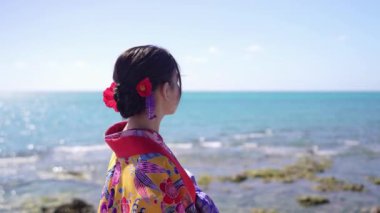 Geleneksel Ryuso ile süslenmiş yirmili yaşlarda genç bir Japon kadın, yavaşça deniz kenarında yürüyor, kültürel zarafetin ve doğal güzelliğin özünü yakalıyor..
