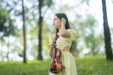 Serin bir fotoğraf, 20 'li yaşlarda, çarpıcı sarı bir elbise giymiş, Kuala Lumpur' da bereketli, doğa dolu bir parkta zarif bir şekilde keman çalan genç bir Çinli-Malezyalı kadını resmediyor.