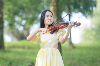 Serin bir fotoğraf, 20 'li yaşlarda, çarpıcı sarı bir elbise giymiş, Kuala Lumpur' da bereketli, doğa dolu bir parkta zarif bir şekilde keman çalan genç bir Çinli-Malezyalı kadını resmediyor.