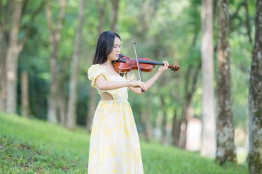 Serin bir fotoğraf, 20 'li yaşlarda, çarpıcı sarı bir elbise giymiş, Kuala Lumpur' da bereketli, doğa dolu bir parkta zarif bir şekilde keman çalan genç bir Çinli-Malezyalı kadını resmediyor.