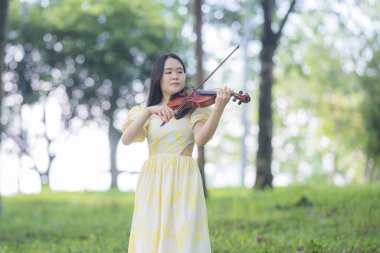 Serin bir fotoğraf, 20 'li yaşlarda, çarpıcı sarı bir elbise giymiş, Kuala Lumpur' da bereketli, doğa dolu bir parkta zarif bir şekilde keman çalan genç bir Çinli-Malezyalı kadını resmediyor.