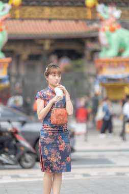 20 'li yaşlarda Tayvanlı genç bir kadın Longshan Tapınağı, Wanhua Bölgesi, Tayvan' ın önünde mavi keongsam takıp köpüklü çay içiyor.