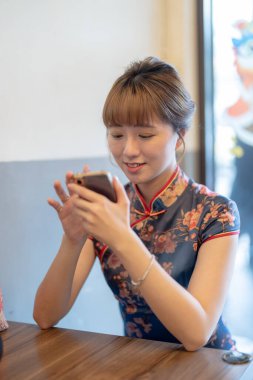 Tayvanlı genç bir kadın, 20 'li yaşlarda mavi bir Cheongsam giyiyor. Wanhua Bölgesi, Tayvan' da bir Çin restoranında yemek siparişini beklerken akıllı telefonunu kullanıyor.