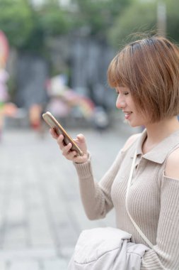 Young Taiwanese woman in her 20s wearing cream-colored clothes, standing and using her smartphone at the famous tourist attraction Longshan Temple in Wanhua District, Taipei, Taiwan clipart