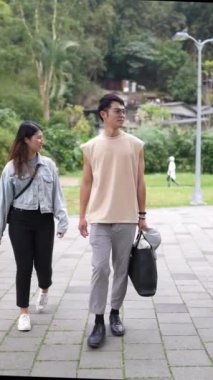 Taipei 'nin Xinyi İlçesi' ndeki bir parkta yirmili yaşlarda genç bir Tayvanlı çiftin bahar öğleden sonra yürüdüğü dikey ağır çekim bir video. Adam kolsuz bir bluz giyiyor.,