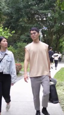 Taipei 'nin Xinyi İlçesi' ndeki bir parkta yirmili yaşlarda genç bir Tayvanlı çiftin bahar öğleden sonra yürüdüğü dikey ağır çekim bir video. Adam kolsuz bir bluz giyiyor.,