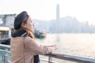 Tsim Sha Tsui, yirmili yaşlarda, kahverengi saçlı, siyah ekose elbiseli, bereli ve mini etekli, Hong Kong 'daki West Kowloon limanının deniz kenarındaki çitlerine yaslanmış bir kadın.