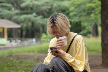Yazın başlarında, Yoyogi Parkı 'nın sessiz bir bölgesinde, Shibuya, Tokyo' da, yirmili yaşlarda, sarı elbiseli, ağaç kütüğünde oturan, kahve içen ve akıllı telefon kullanan bir Japon..