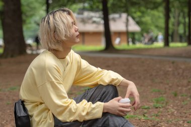 Yazın başlarında, Yoyogi Parkı 'nın sessiz bir bölgesinde, Shibuya, Tokyo' da, yirmili yaşlarda, sarı elbiseli, ağaç kütüğünde oturan, kahve içen ve akıllı telefon kullanan bir Japon..