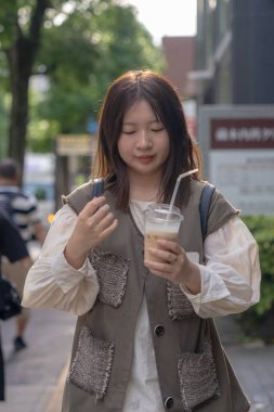 Yazın başlarında, genç bir Çinli değişim öğrencisi, 20 'li yaşlarda, Tokyo, Minato' daki Hamamatsucho İstasyonu 'nun önünde elinde plastik bir bardakta buzlu bir au lait tutuyor..