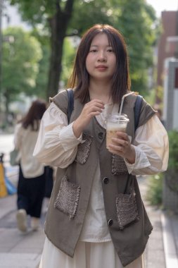 Yazın başlarında, genç bir Çinli değişim öğrencisi, 20 'li yaşlarda, Tokyo, Minato' daki Hamamatsucho İstasyonu 'nun önünde elinde plastik bir bardakta buzlu bir au lait tutuyor..