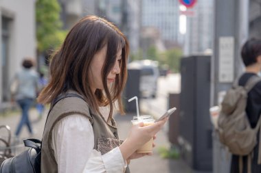 Yazın başlarında, genç bir Çinli değişim öğrencisi, 20 'li yaşlarda, Tokyo, Minato' daki Hamamatsucho İstasyonu 'nun önünde elinde plastik bir bardakta buzlu bir au lait tutuyor..