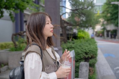 Yazın başlarında, genç bir Çinli değişim öğrencisi, 20 'li yaşlarda, Tokyo, Minato' daki Hamamatsucho İstasyonu 'nun önünde elinde plastik bir bardakta buzlu bir au lait tutuyor..