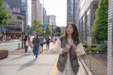 Yazın başlarında, genç bir Çinli değişim öğrencisi, 20 'li yaşlarda, Tokyo, Minato' daki Hamamatsucho İstasyonu 'nun önünde elinde plastik bir bardakta buzlu bir au lait tutuyor..
