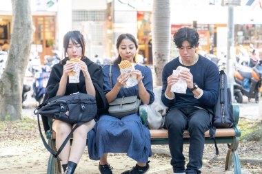 Tayvanlı bir kadın, Tayvanlı bir erkek ve Asyalı bir kadın, Taipei 'de Daan Bölgesi' nde bir bankta oturmuş, büyük bir waffle 'ın keyfini çıkarıyorlar..