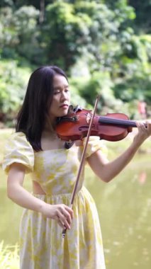 Malezya 'nın Kuala Lumpur kenti yakınlarındaki Bukit Jalil' de, büyük doğa dolu bir parkta, göl kenarında yavaşça yürüyen 20 'li yaşlarda, sarı elbiseli Çinli bir kadın. (yavaş çekim dikey video)