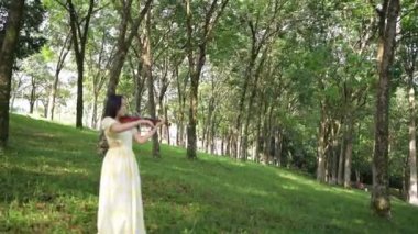 20 'li yaşlarda, sarı elbiseli Çinli bir kadın Malezya, Kuala Lumpur yakınlarındaki Bukit Jalil' deki büyük doğa dolu bir parkta keman çalıyor..