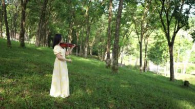 20 'li yaşlarda, sarı elbiseli Çinli bir kadın Malezya, Kuala Lumpur yakınlarındaki Bukit Jalil' deki büyük doğa dolu bir parkta keman çalıyor..