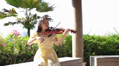 20 'li yaşlarda, sarı elbiseli Çinli bir kadın Malezya' nın Kuala Lumpur yakınlarındaki Bukit Jalil 'de yeşillik ve çiçeklerle çevrili bir çardakta keman çalıyor..