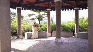 20 'li yaşlarda, sarı elbiseli Çinli bir kadın Malezya' nın Kuala Lumpur yakınlarındaki Bukit Jalil 'de yeşillik ve çiçeklerle çevrili bir çardakta keman çalıyor..