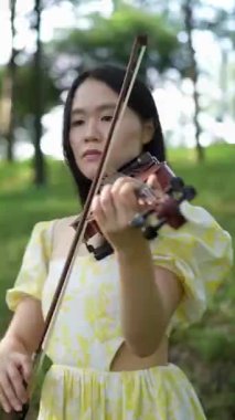20 'li yaşlarda, sarı elbiseli Çinli bir kadın Malezya, Kuala Lumpur yakınlarındaki Bukit Jalil' deki büyük doğa dolu bir parkta keman çalıyor. (dikey video)