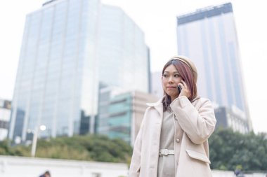 Hong Kong 'lu, kızılımsı kahverengi saçlı bir kadın akıllı telefonuyla konuşuyor. Kowloon Park, Tsim Sha Tsui, Kowloon Adası, Hong Kong' daki yüksek bina kümesinin arka planında..