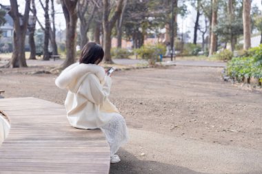 20 'li yaşlarda, siyah saçlı, beyaz önlüklü, tüylü, beyaz etekli bir kadın parkta oturuyor ve Minato, Tokyo, Japonya' da akıllı telefonunu kullanıyor..