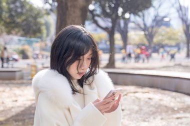 20 'li yaşlarda, siyah saçlı, beyaz önlüklü, tüylü, beyaz etekli bir kadın parkta oturuyor ve Minato, Tokyo, Japonya' da akıllı telefonunu kullanıyor..