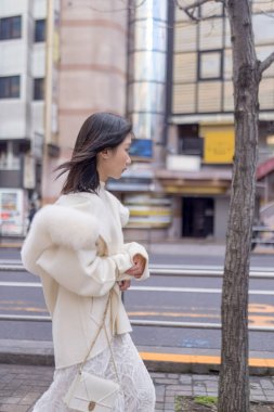 20 'li yaşlarda, siyah saçlı, beyaz önlüklü, tüylü ve beyaz etekli bir Çinli kadın. Minato, Tokyo, Japonya' da halka açık bir yolda yürüyor..