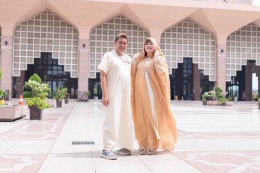50 'li yaşlardaki Müslüman Malezyalı, beyaz giysili, siyah şapkalı ve krem renkli Çinli kadın Putrajaya, Malezya' da büyük bir cami önünde yan yana duruyor..