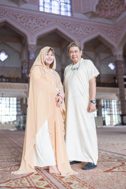 50-something Muslim Malay man in white outfit and black hat and 30-something Chinese woman in cream-colored religious attire standing together inside large mosque in Putrajaya, Malaysia. clipart