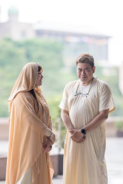 50-something Muslim Malay man in white outfit and black hat and 30-something Chinese woman in cream-colored religious attire standing together outside large mosque in Putrajaya, Malaysia. clipart