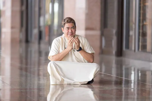 stock image Fifty-something Muslim Malay person in religious attire, wearing a white outfit and black hat, praying devoutly in a wide glass-enclosed marble area outside very large mosque in Putrajaya, Malaysia.