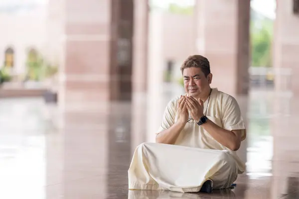 Stock image Fifty-something Muslim Malay person in religious attire, wearing a white outfit and black hat, praying devoutly in a wide glass-enclosed marble area outside very large mosque in Putrajaya, Malaysia.