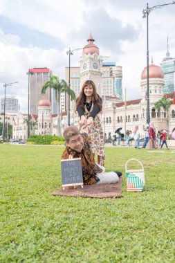 50 'li yaşlarda Malezyalı bir erkek ve 30' lu yaşlarda bir Çinli kadın Malezya 'nın başkenti Kuala Lumpur' da bulunan tarihi binanın önünde karatahtayla fotoğraf çekiyorlar..