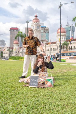 50 'li yaşlarda Malezyalı bir erkek ve 30' lu yaşlarda bir Çinli kadın Malezya 'nın başkenti Kuala Lumpur' da bulunan tarihi binanın önünde karatahtayla fotoğraf çekiyorlar..