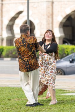 50 'li yaşlarda Malezyalı bir adam ve 30' lu yaşlarda Çinli bir kadın, ikisi de geleneksel Malezya kıyafeti giymiş, Malezya 'nın başkenti Kuala Lumpur' da tarihi bir binanın önünde dans ediyorlar..