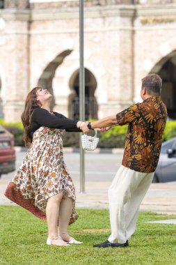 50 'li yaşlarda Malezyalı bir adam ve 30' lu yaşlarda Çinli bir kadın, ikisi de geleneksel Malezya kıyafeti giymiş, Malezya 'nın başkenti Kuala Lumpur' da tarihi bir binanın önünde dans ediyorlar..