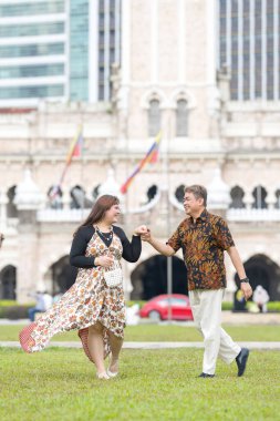 50 'li yaşlarda Malezyalı bir adam ve 30' lu yaşlarda Çinli bir kadın, ikisi de geleneksel Malezya kıyafeti giymiş, Malezya 'nın başkenti Kuala Lumpur' da tarihi bir binanın önünde dans ediyorlar..