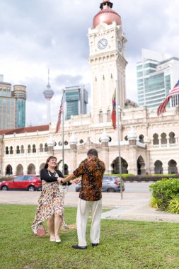 50 'li yaşlarda Malezyalı bir adam ve 30' lu yaşlarda Çinli bir kadın, ikisi de geleneksel Malezya kıyafeti giymiş, Malezya 'nın başkenti Kuala Lumpur' da tarihi bir binanın önünde dans ediyorlar..