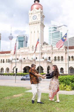 50 'li yaşlarda Malezyalı bir adam ve 30' lu yaşlarda Çinli bir kadın, ikisi de geleneksel Malezya kıyafeti giymiş, Malezya 'nın başkenti Kuala Lumpur' da tarihi bir binanın önünde dans ediyorlar..