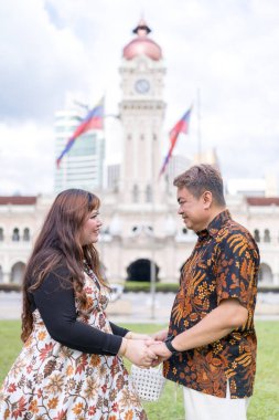 50 'li yaşlarda Malezyalı bir adam ve 30' lu yaşlarda Çinli bir kadın. İkisi de geleneksel Malezya kıyafeti giymiş. Malezya 'nın başkenti Kuala Lumpur' daki tarihi binanın önünde birbirlerinin gözlerinin içine bakıyor..