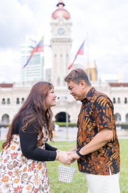 50 'li yaşlarda Malezyalı bir adam ve 30' lu yaşlarda Çinli bir kadın. İkisi de geleneksel Malezya kıyafeti giymiş. Malezya 'nın başkenti Kuala Lumpur' daki tarihi binanın önünde birbirlerinin gözlerinin içine bakıyor..