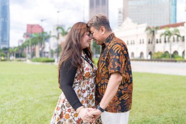 50 'li yaşlarda Malezyalı bir adam ve 30' lu yaşlarda Çinli bir kadın. İkisi de geleneksel Malezya kıyafeti giymiş. Malezya 'nın başkenti Kuala Lumpur' daki tarihi binanın önünde birbirlerinin gözlerinin içine bakıyor..