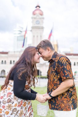 50 'li yaşlarda Malezyalı bir adam ve 30' lu yaşlarda Çinli bir kadın. İkisi de geleneksel Malezya kıyafeti giymiş. Malezya 'nın başkenti Kuala Lumpur' daki tarihi binanın önünde birbirlerinin gözlerinin içine bakıyor..