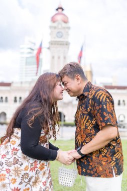 50 'li yaşlarda Malezyalı bir adam ve 30' lu yaşlarda Çinli bir kadın. İkisi de geleneksel Malezya kıyafeti giymiş. Malezya 'nın başkenti Kuala Lumpur' daki tarihi binanın önünde birbirlerinin gözlerinin içine bakıyor..