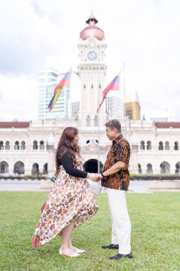 50 'li yaşlarda Malezyalı bir adam ve 30' lu yaşlarda Çinli bir kadın. İkisi de geleneksel Malezya kıyafeti giymiş. Malezya 'nın başkenti Kuala Lumpur' daki tarihi binanın önünde birbirlerinin gözlerinin içine bakıyor..