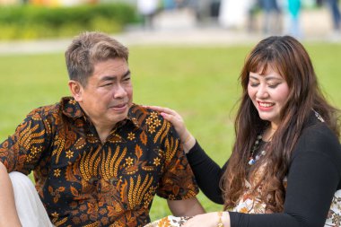 50 'li yaşlarda Malezyalı bir adam ve 30' lu yaşlarda Çinli bir kadın. İkisi de Malezya 'nın başkenti Kuala Lumpur' da bulunan tarihi bir binanın önündeki çimlerin üzerinde oturuyorlar..