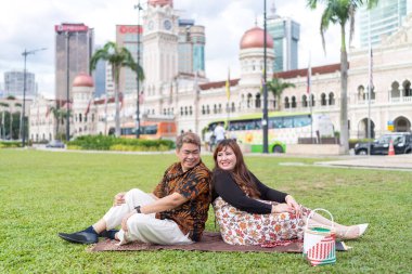 50 'li yaşlarda Malezyalı bir adam ve 30' lu yaşlarda Çinli bir kadın. İkisi de Malezya 'nın başkenti Kuala Lumpur' da bulunan tarihi bir binanın önündeki çimlerin üzerinde oturuyorlar..