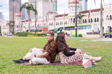 50 'li yaşlarda Malezyalı bir adam ve 30' lu yaşlarda Çinli bir kadın. İkisi de Malezya 'nın başkenti Kuala Lumpur' da bulunan tarihi bir binanın önündeki çimlerin üzerinde oturuyorlar..